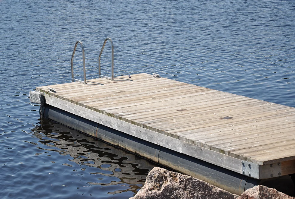 A wooden dock with a metal ladder extends over a calm, blue body of water, gently lifting above the tidal flow. Large rocks are visible in the foreground, framing the dock. The scene is peaceful and serene.