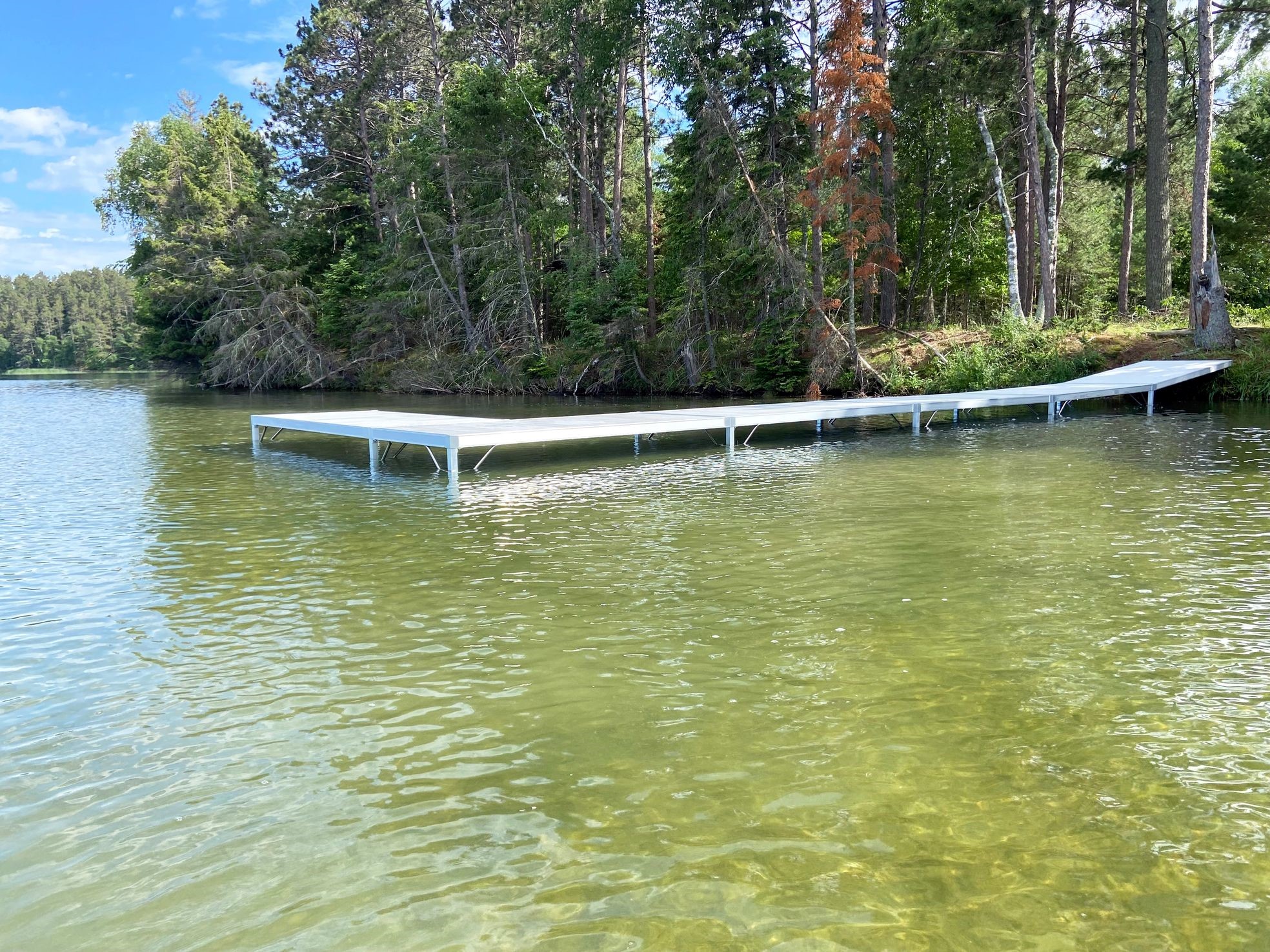 A scenic view of a wooden dock extending over clear, shallow water in a tranquil lake, where the gentle tidal lifts harmoniously with nature, surrounded by lush green trees under a bright blue sky with scattered clouds.