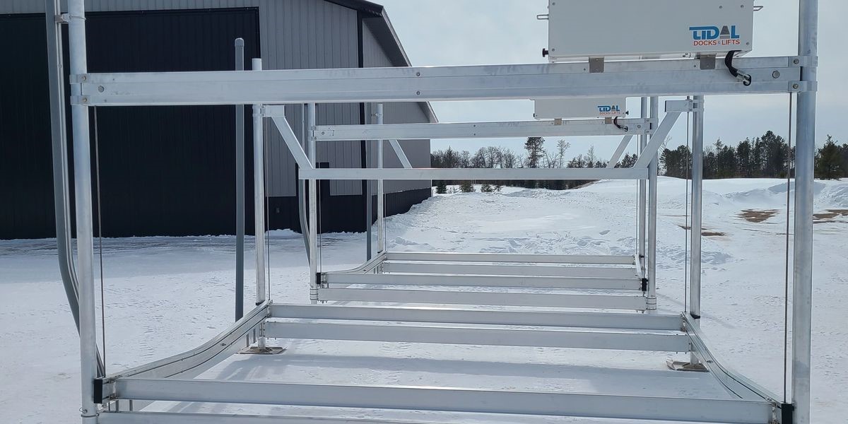 Metal boat lift frames are stacked in the snow near a large black building with a sign reading "Tidal Dock Lifts." Providing excellent service, snow covers the ground, and trees stand visible in the distance under a cloudy sky.