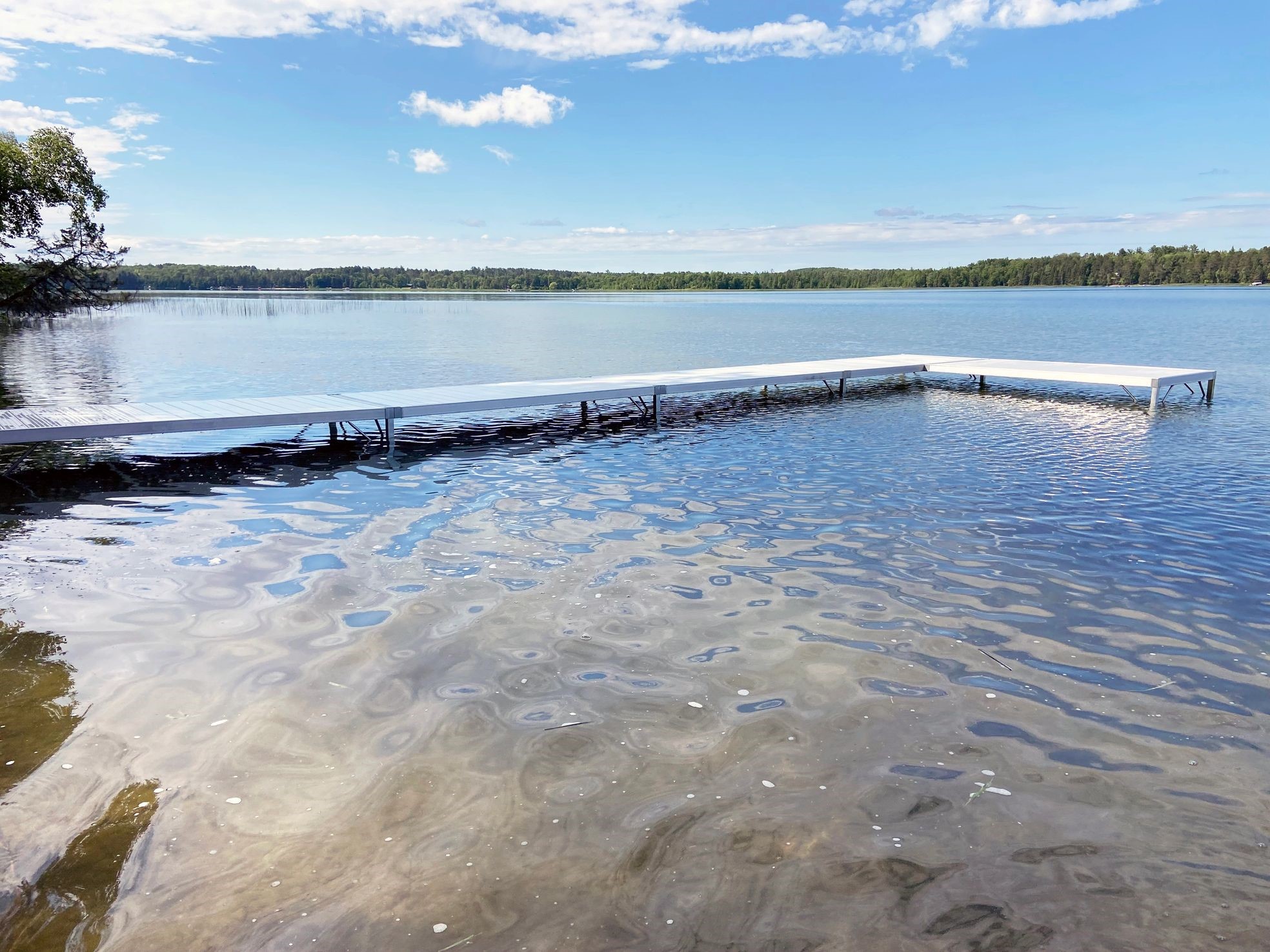 A calm lake with a long dock gracefully extending into the water on a clear, sunny day. The sky is partly cloudy, and trees line the far shore, providing a picturesque service to nature’s beauty.