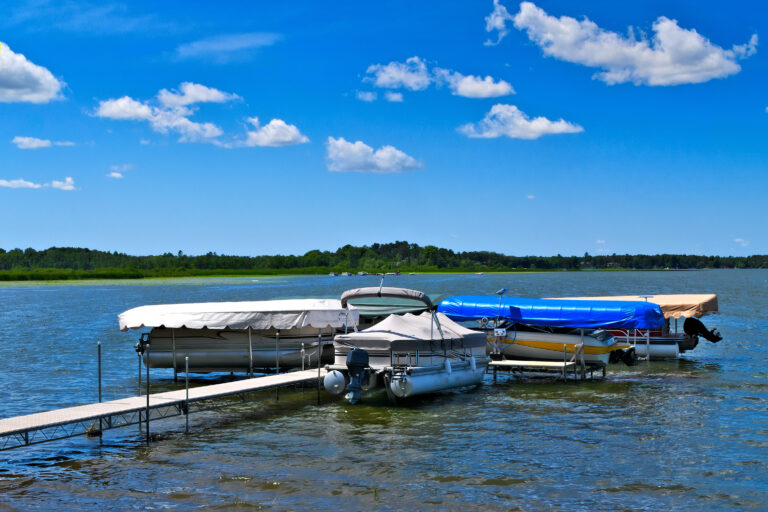 Several boats, including a service barge, are docked on a calm lake under a bright blue sky with scattered clouds. A wooden pier extends into the water, and the shoreline is visible in the distance with lifts of green trees.