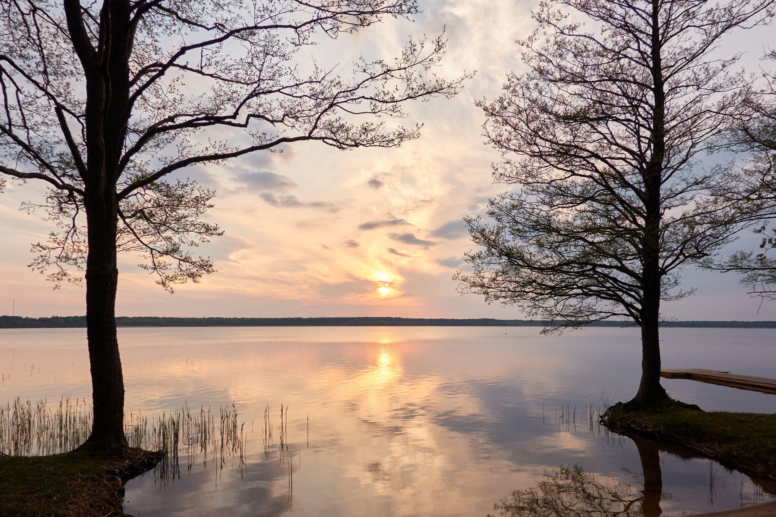 A calm lake at sunset with the sun low in the sky, reflecting on the water. Silhouetted trees frame a peaceful boat lift and dock maintenance area, while sparse clouds scatter across the pastel-colored sky.