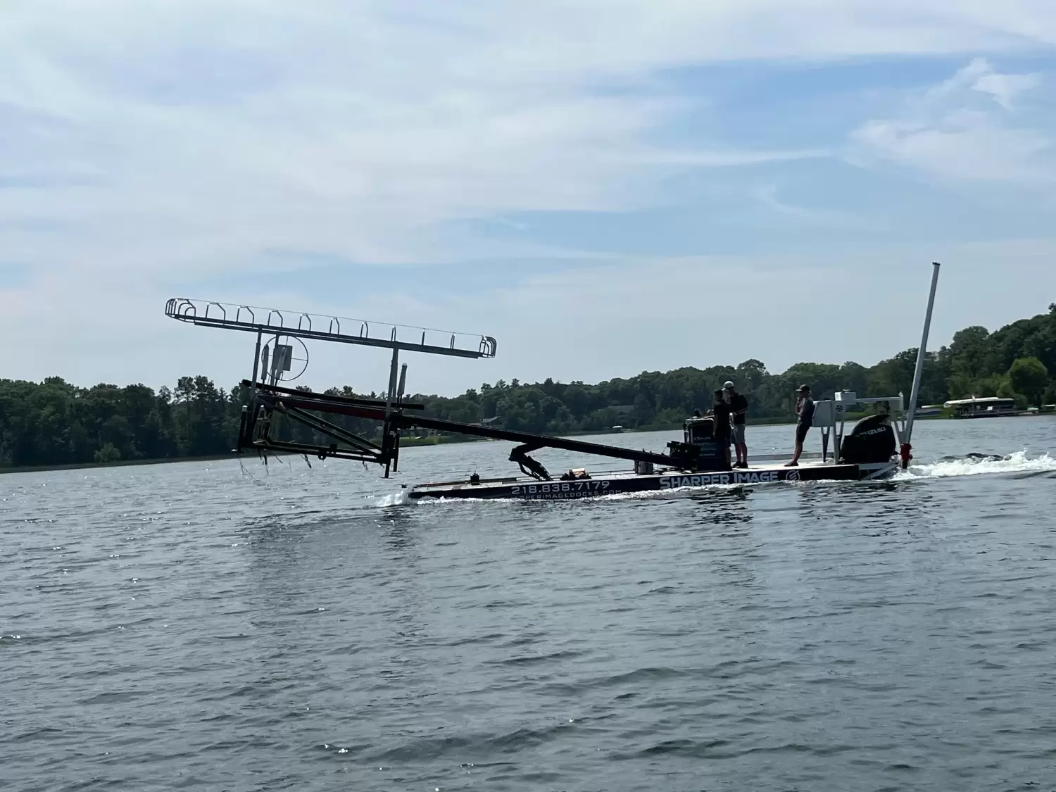 A large black boat with a raised metal structure on its deck, possibly equipped with simple dock accessories, moves across a calm lake with two people on board. Trees line the distant shore under a partly cloudy sky.