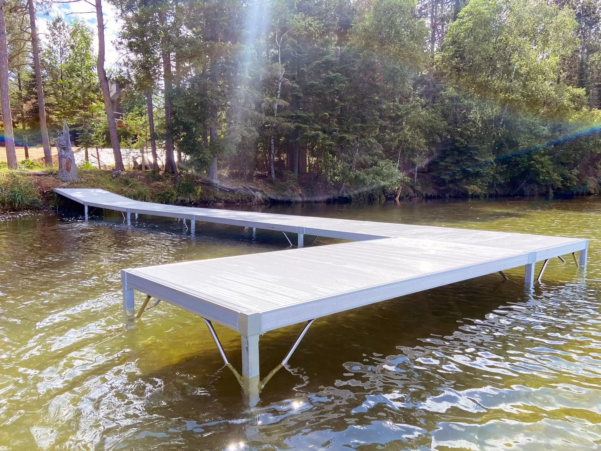 A modern, light-colored dock with metal legs extends over clear, shallow lake water toward a forested shoreline on a sunny day, reflecting careful lake dock maintenance and long-term dock performance. Sunlight creates a visible lens flare in the image.