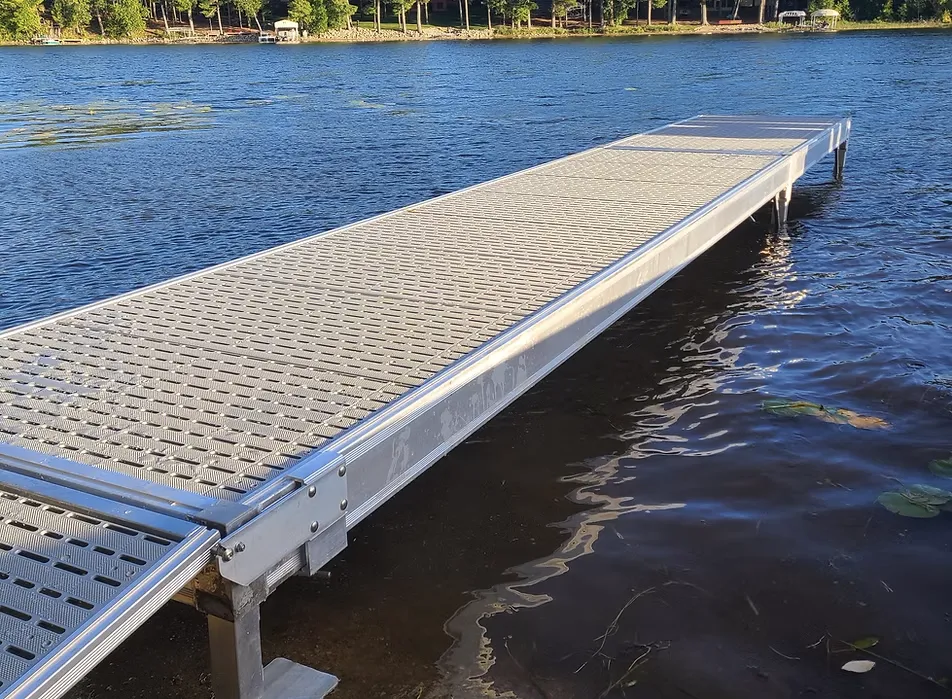A metal dock extends over the edge of a calm blue lake, with sturdy docks and lifts nearby, framed by trees and grassy shorelines glowing under the sunlight.