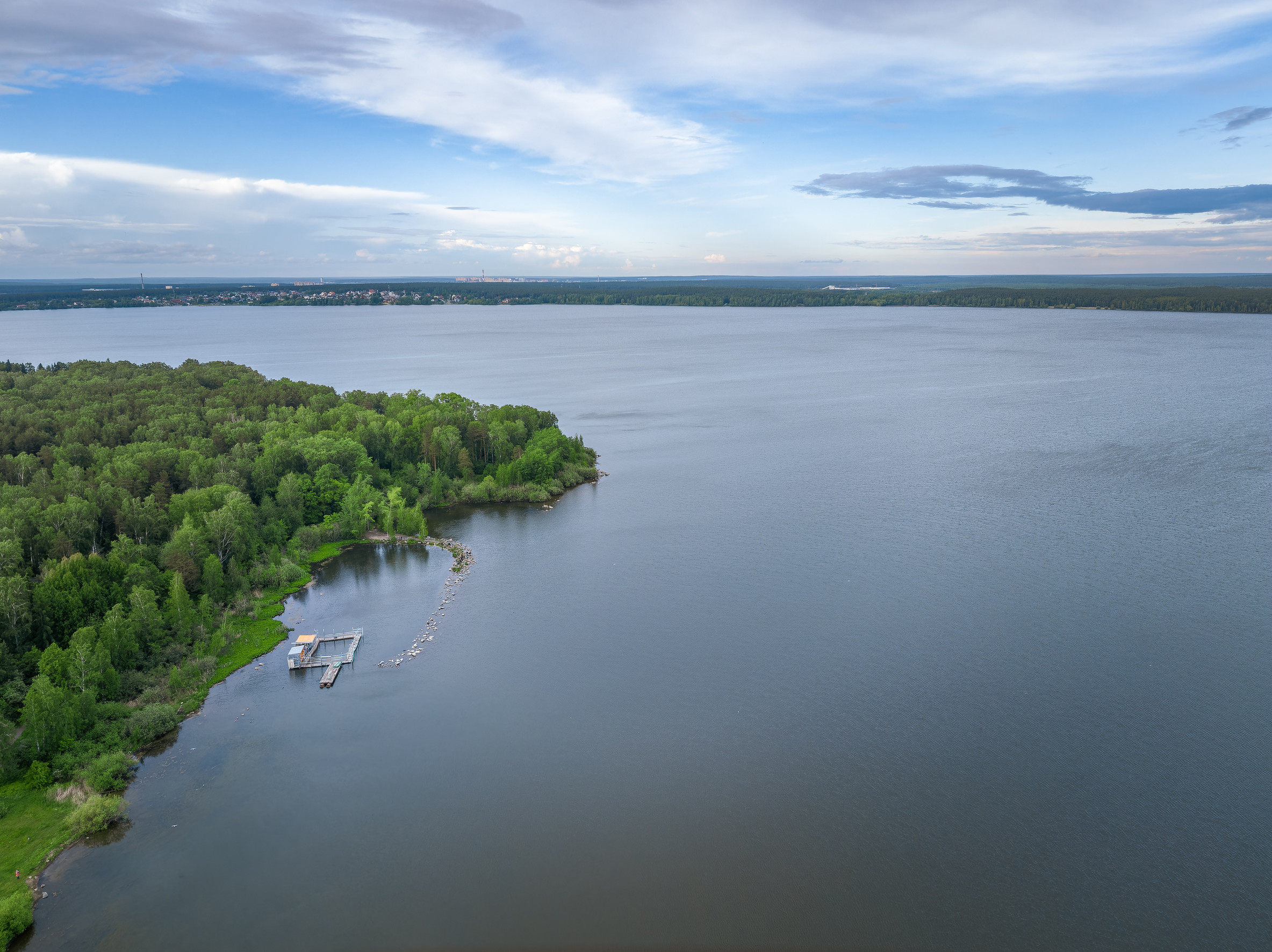 Aerial view of a calm lake with lakefront docks and boats near a green, forested shoreline, under a partly cloudy blue sky—enhance your lakefront with Sidco Docks.