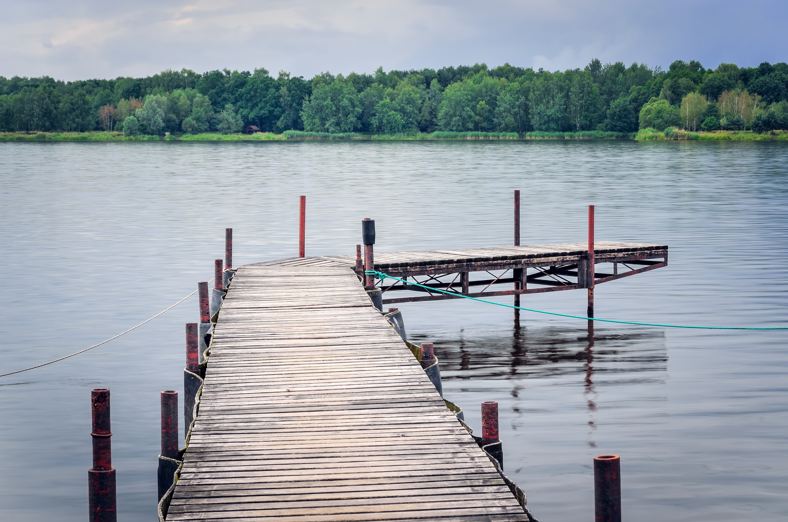A wooden dock extends over a calm lake, with a smaller dock branching off to the right. Trees line the far shore under a cloudy sky, and the water gently reflects the surroundings.