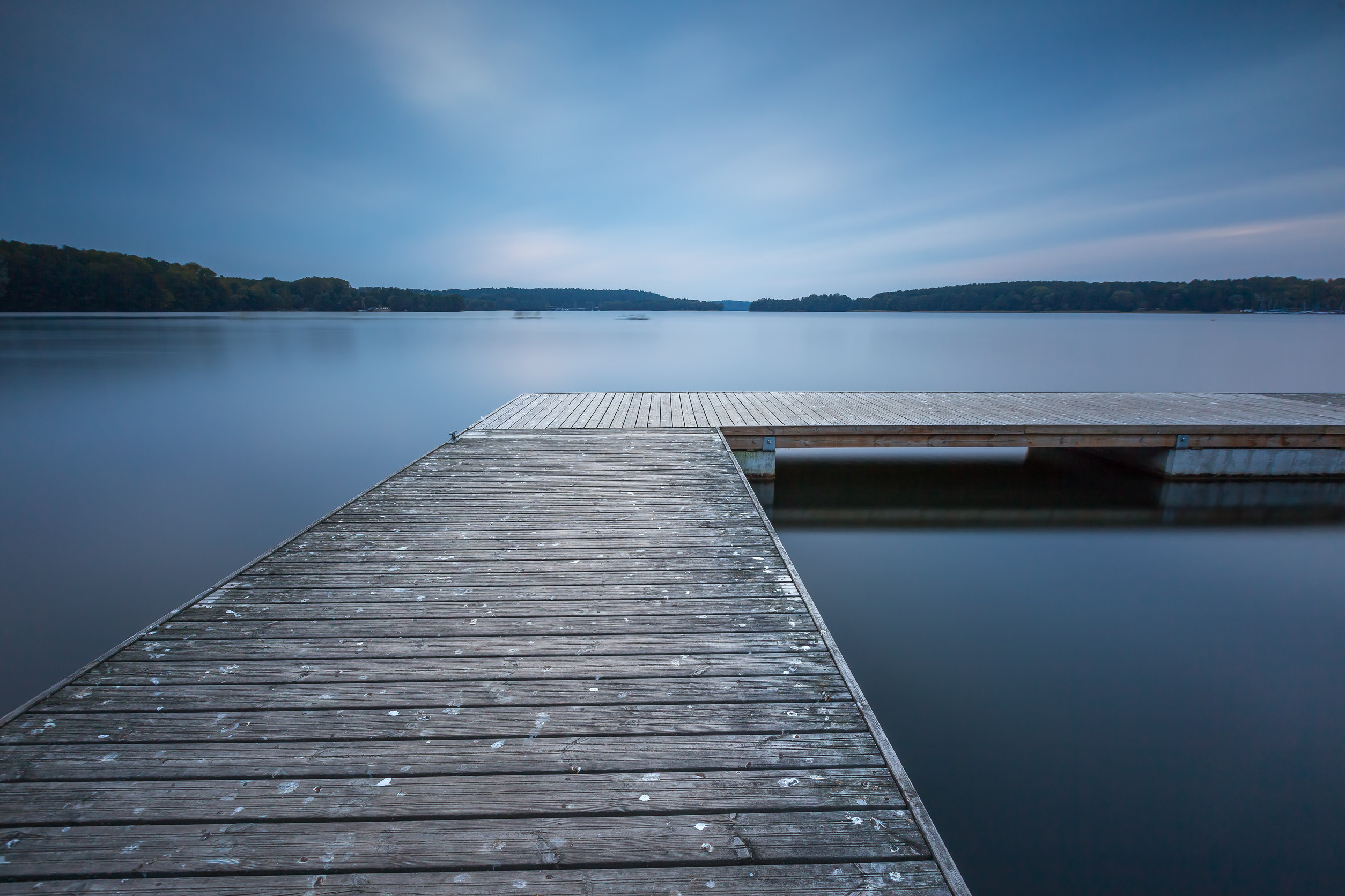 A wooden dock extends over calm, glassy water under a blue sky, with tree-lined shores visible in the distance. The scene is peaceful and serene, suggesting early morning or evening.