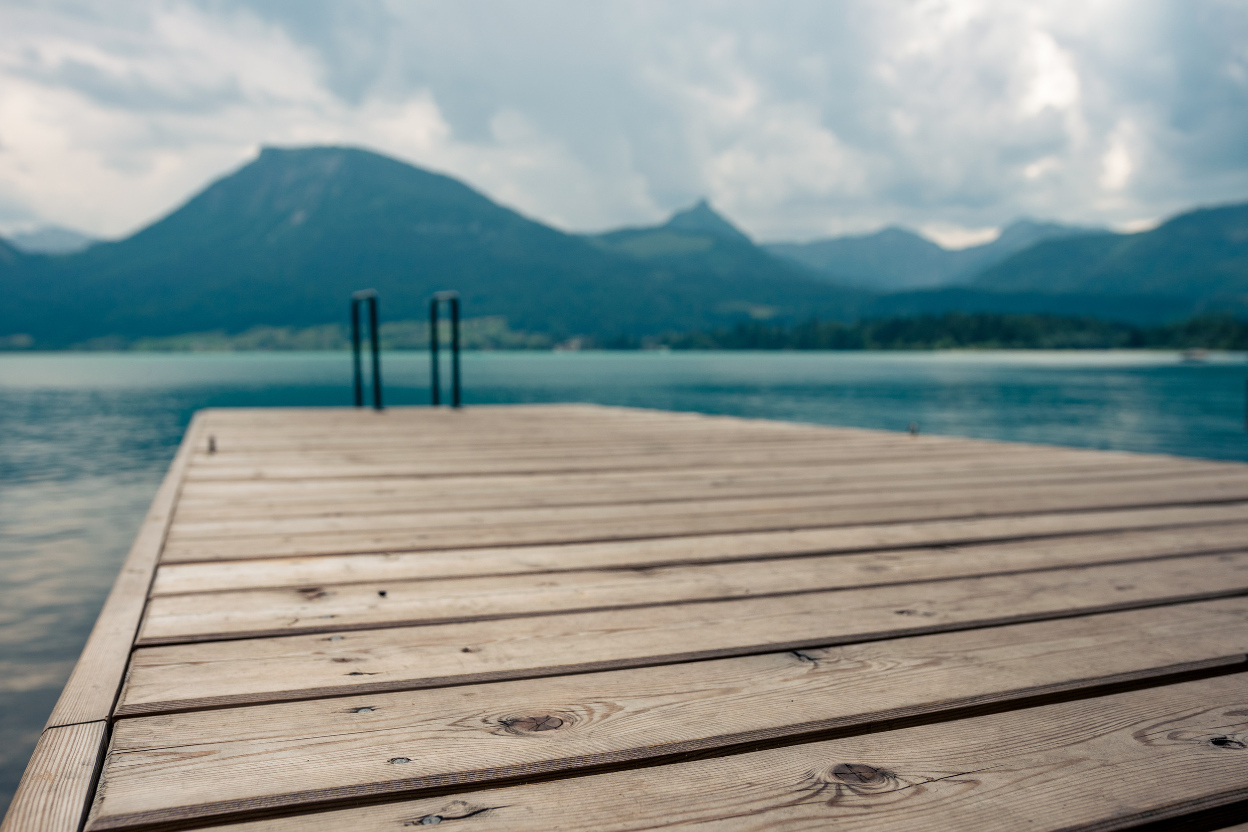 A wooden dock extends over calm water, with metal railings at the end. In the background, there are mountains under a cloudy sky. The scene is peaceful and serene.