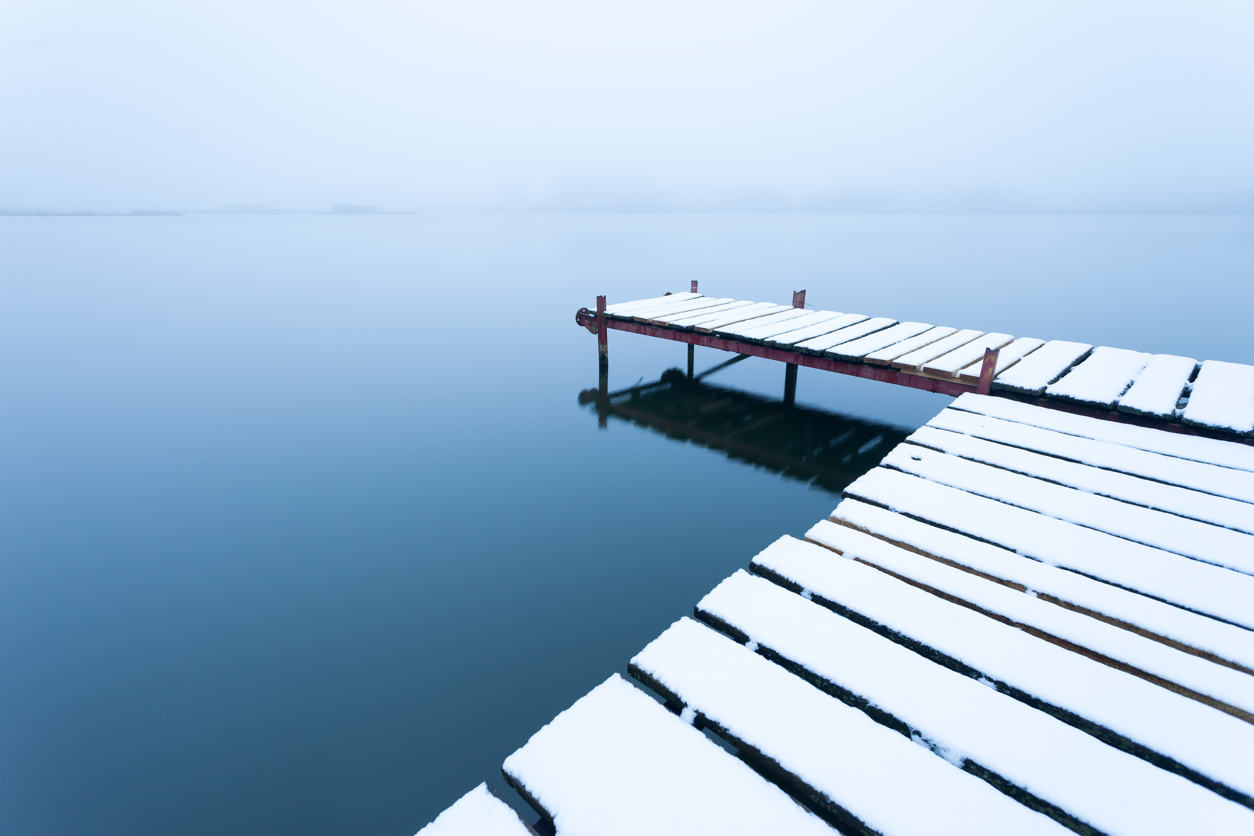 A snow-covered Minnesota Dock extends over a calm, misty lake. The water reflects the soft, pale sky, creating a serene and tranquil winter scene with minimal visibility in the background.