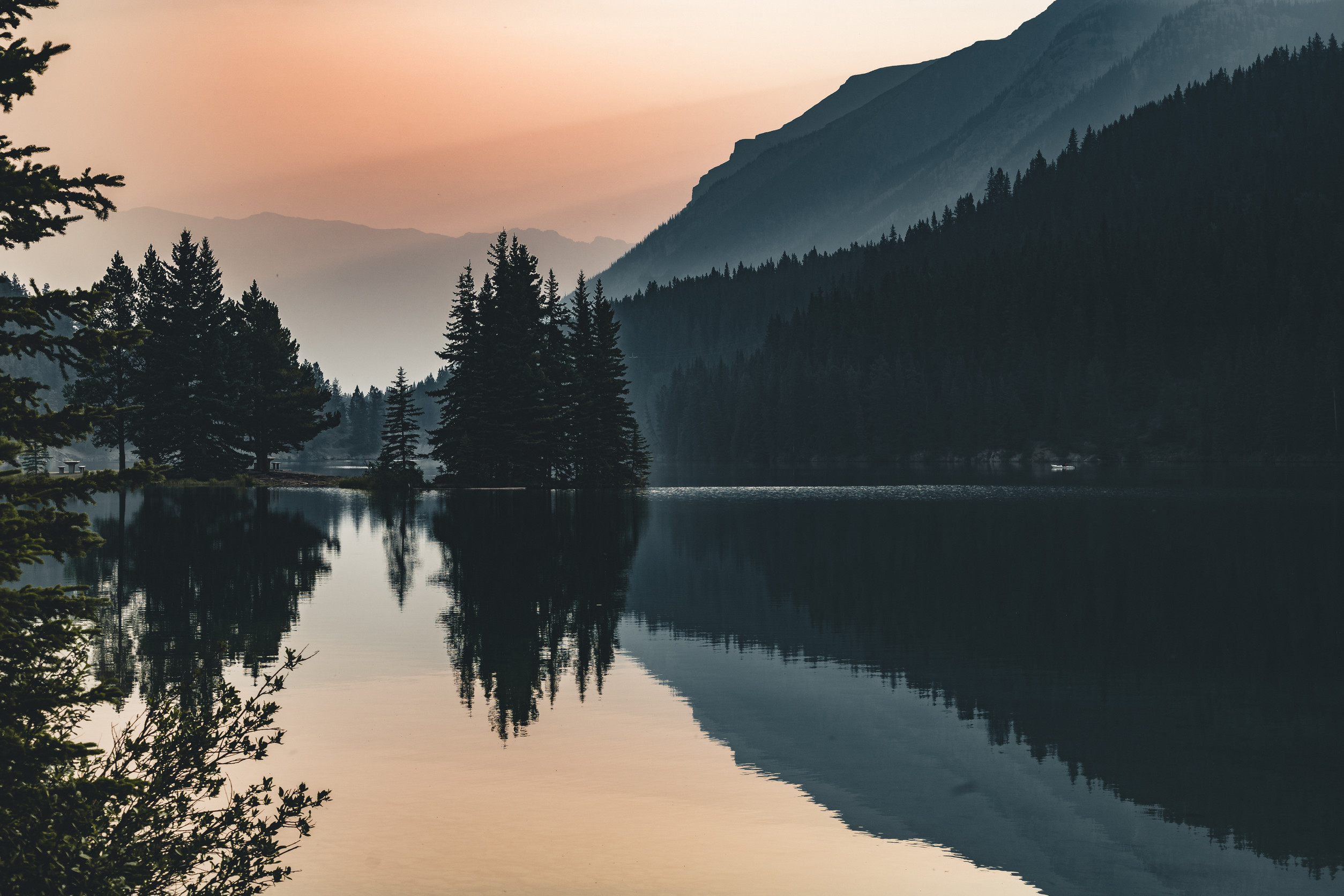 A tranquil lake reflects pine trees and distant mountains at sunset, with soft pastel colors in the sky and mist creating a calm, serene atmosphere.