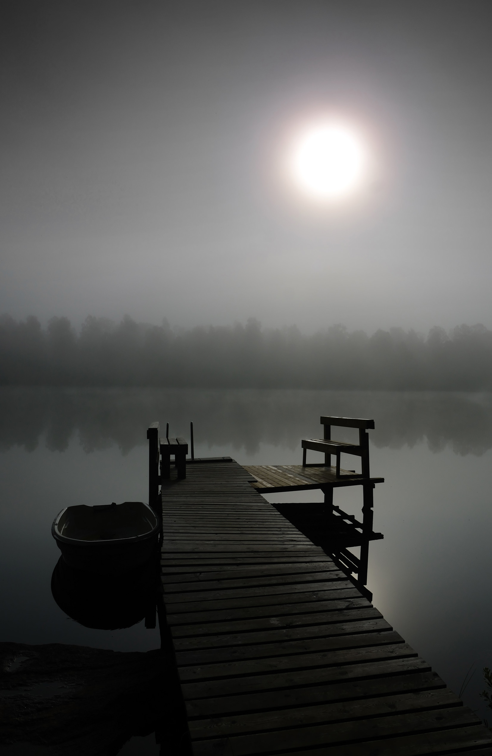A wooden dock extends over a still lake, with a rowboat tied alongside. The scene is shrouded in mist, and a bright sun glows through the fog, silhouetting trees along the distant shoreline.