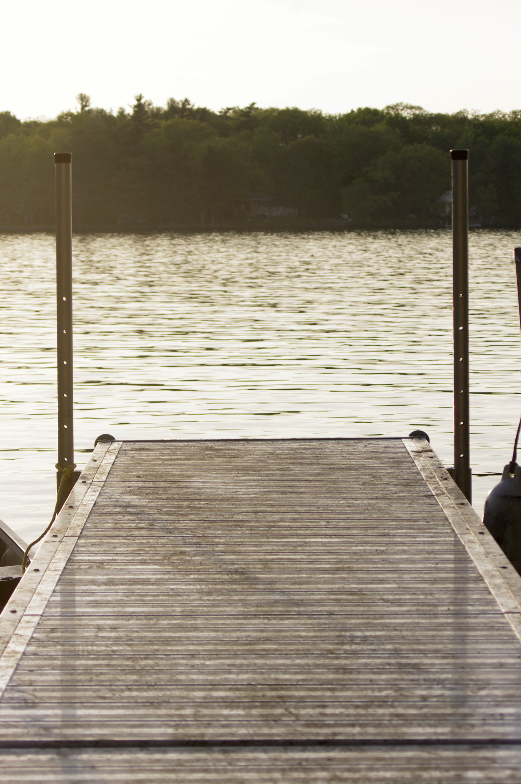 A wooden dock leads out over calm, reflective water with trees lining the opposite shore in the background on a hazy day.
