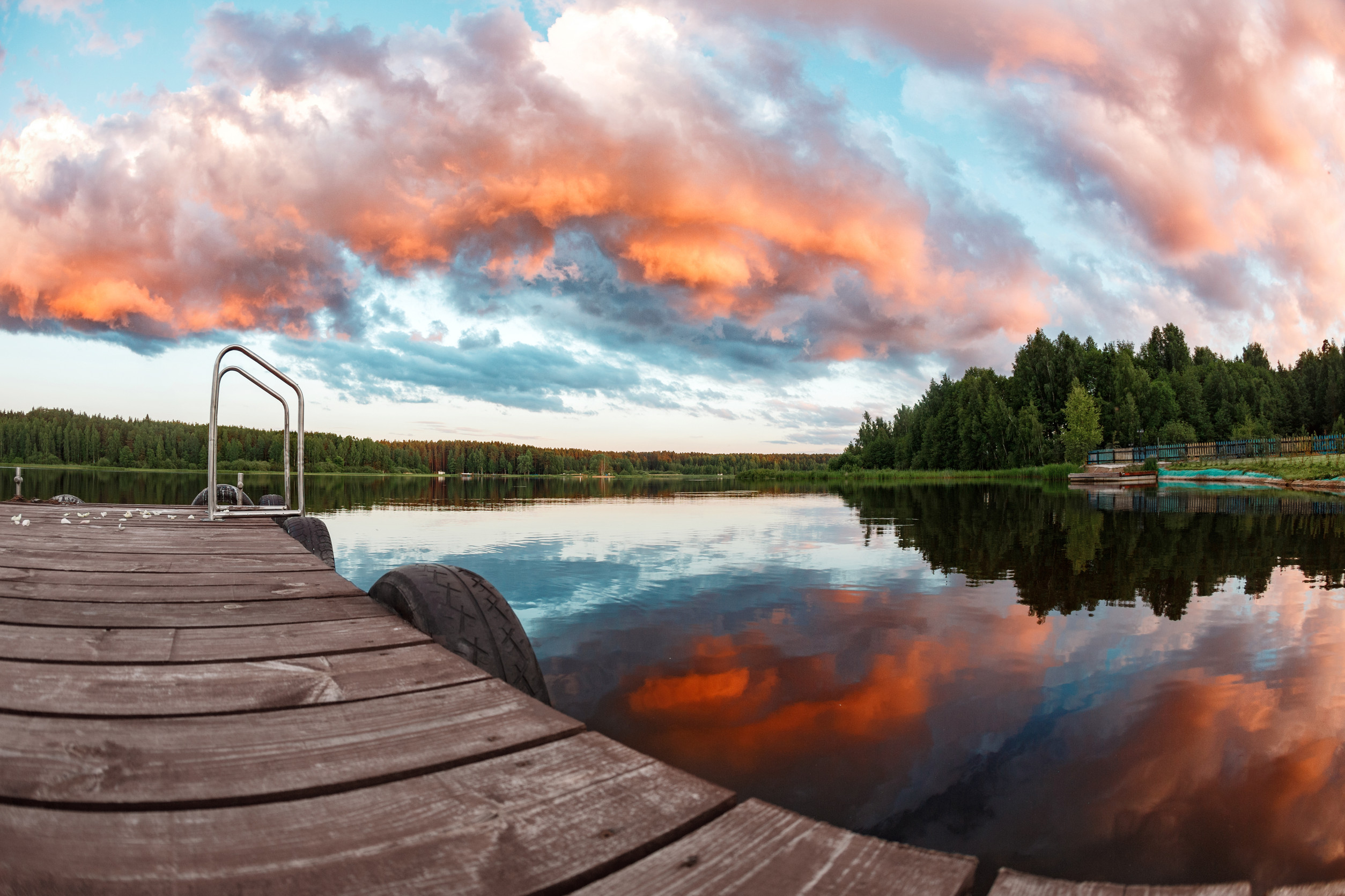 A Minnesota dock extends over a calm lake reflecting dramatic orange and pink clouds at sunset, with a forested shoreline and a distant metal ladder on the left side.
