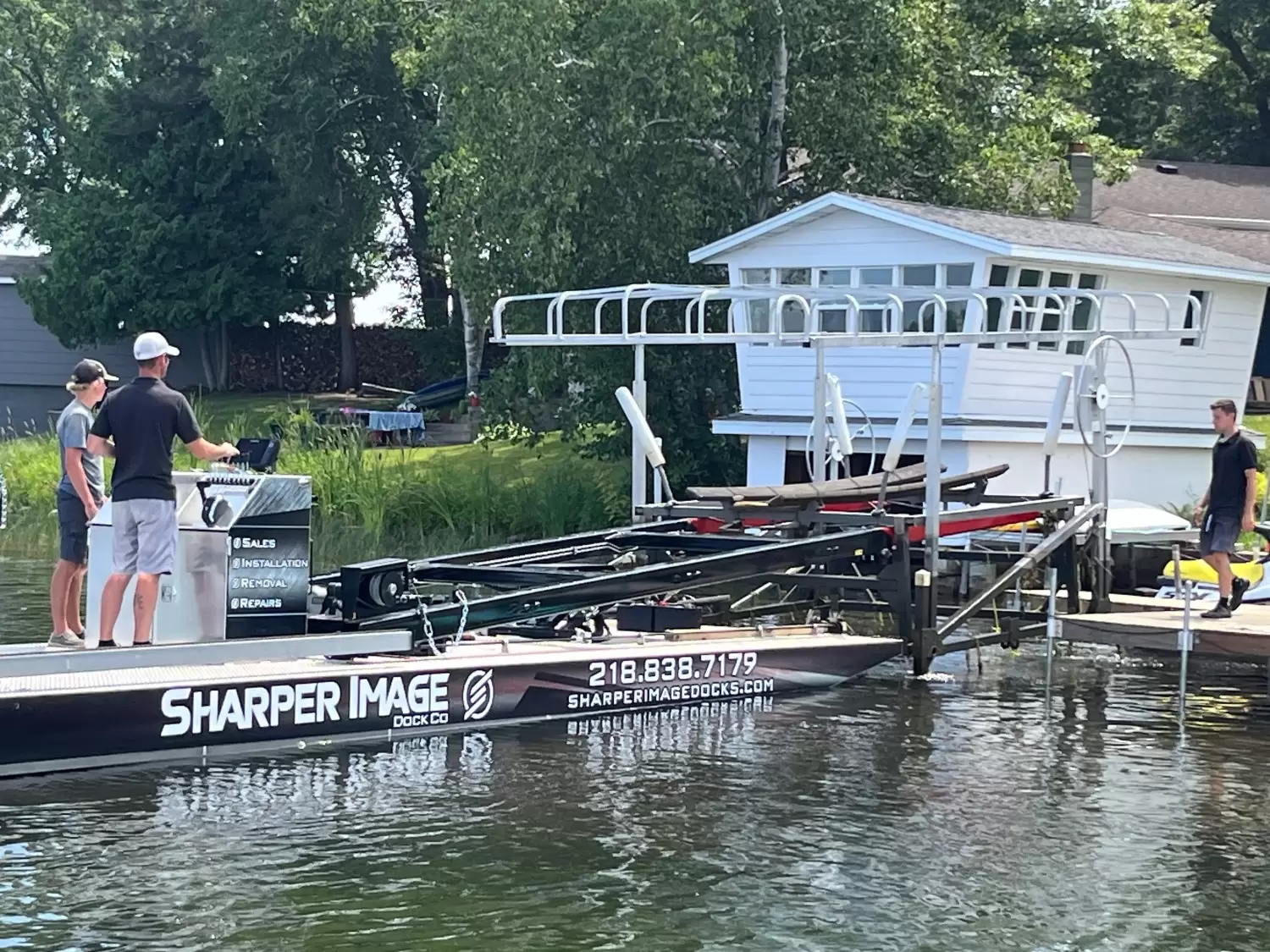 Three people stand on a docked Sharper Image Dock & Lift boat, equipped for spring dock installation, near a lakefront house surrounded by lush greenery.