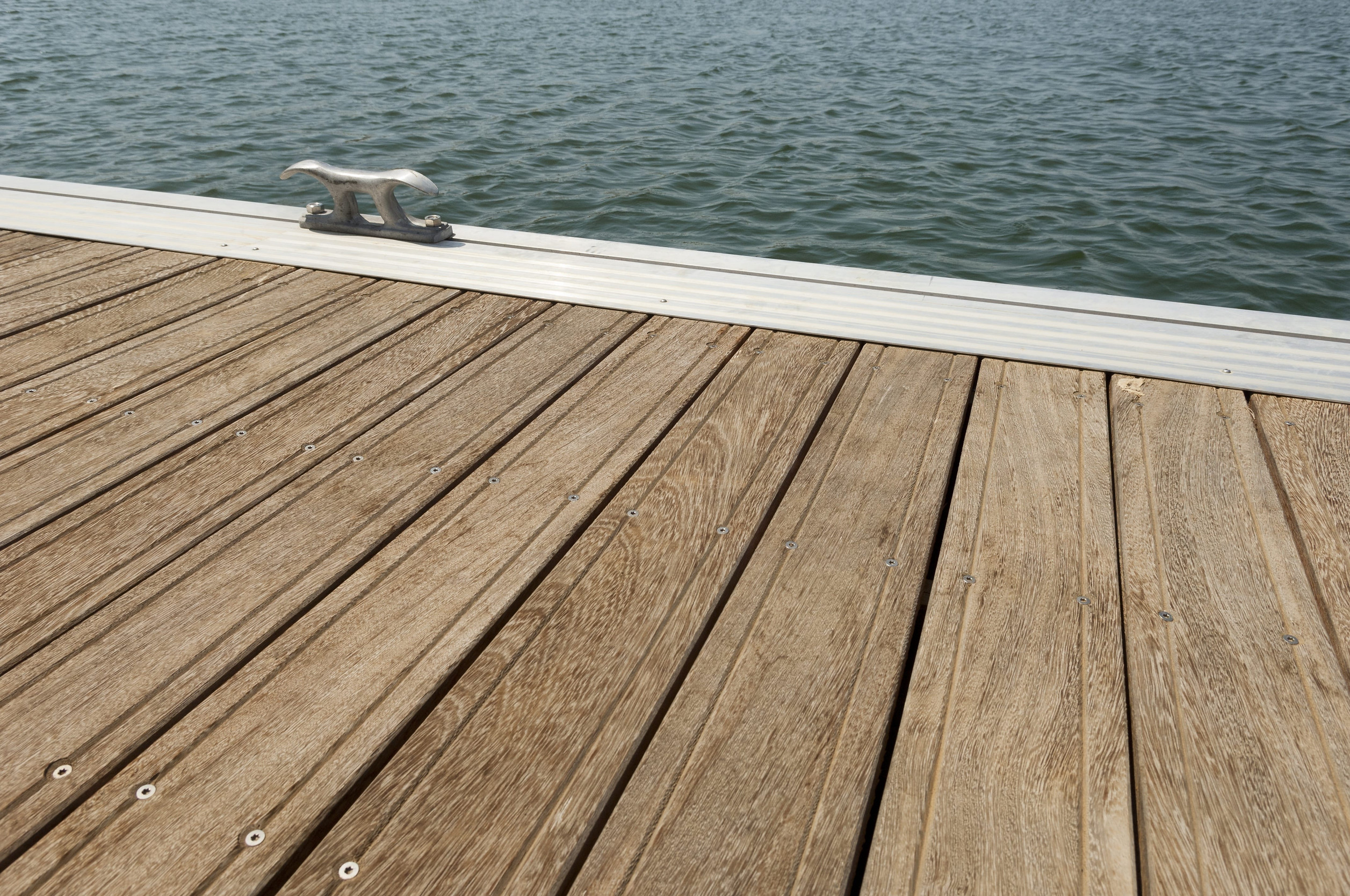 A close-up view of a wooden dock with a metal cleat beside calm, rippling lake water on a serene spring day.