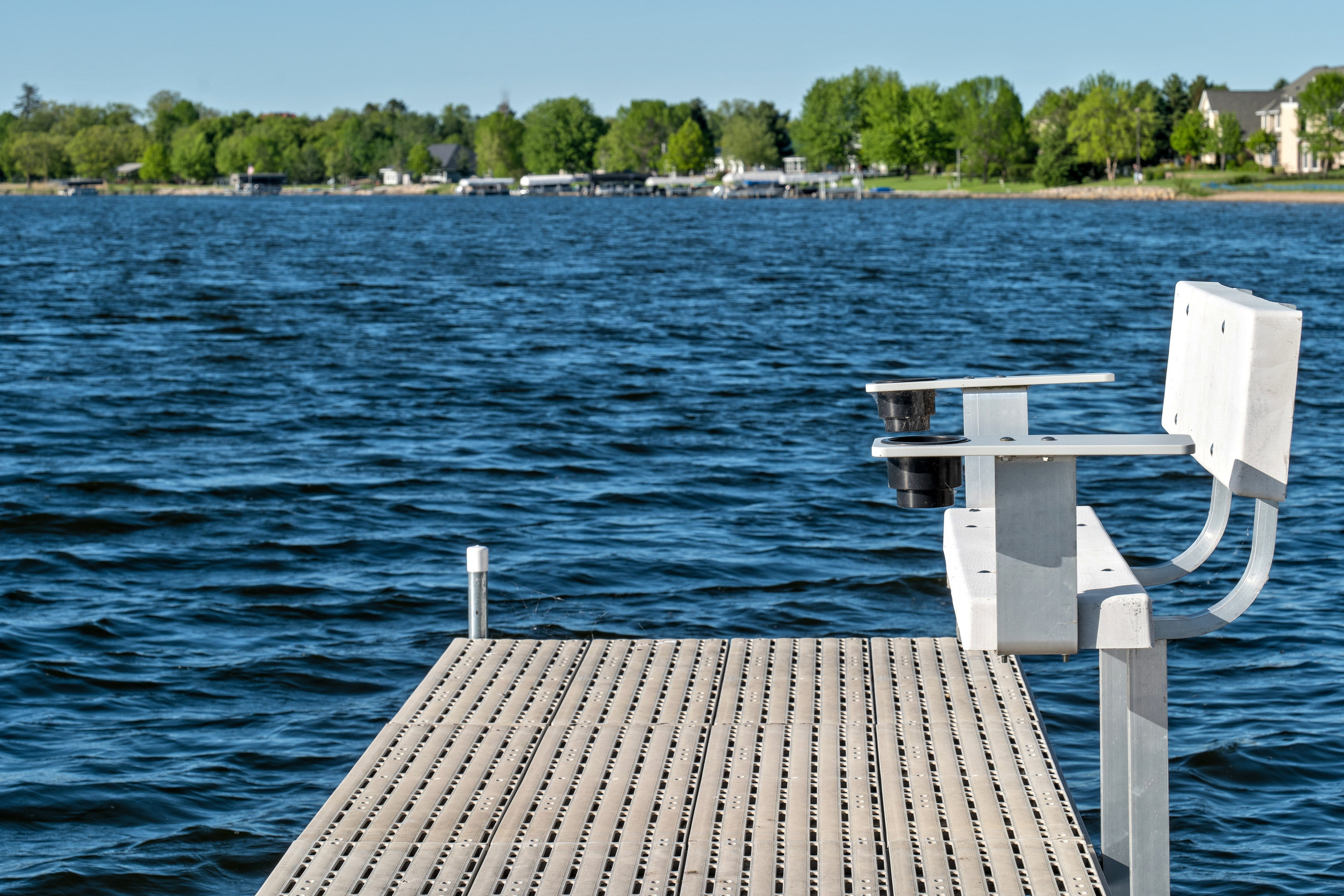 A wooden, stronger dock with a white bench extends over a blue lake. Trees and houses line the distant shore under a clear sky, creating a peaceful and secure waterside scene.