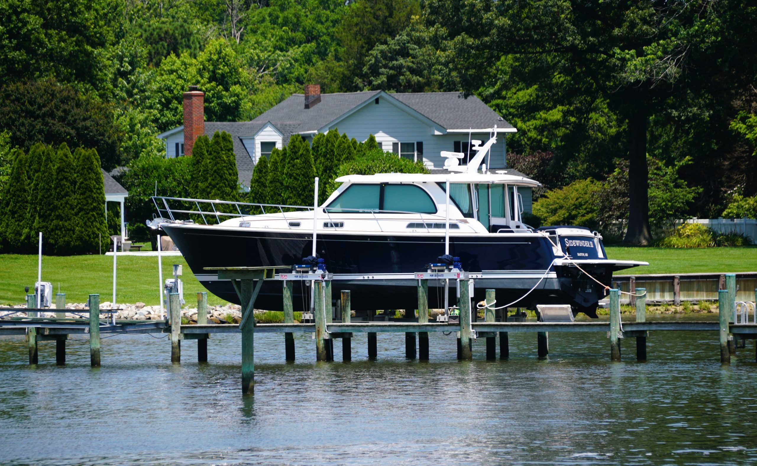Motor yacht on a lift at a dock beside a grassy yard and tall trees, with a house in the background.