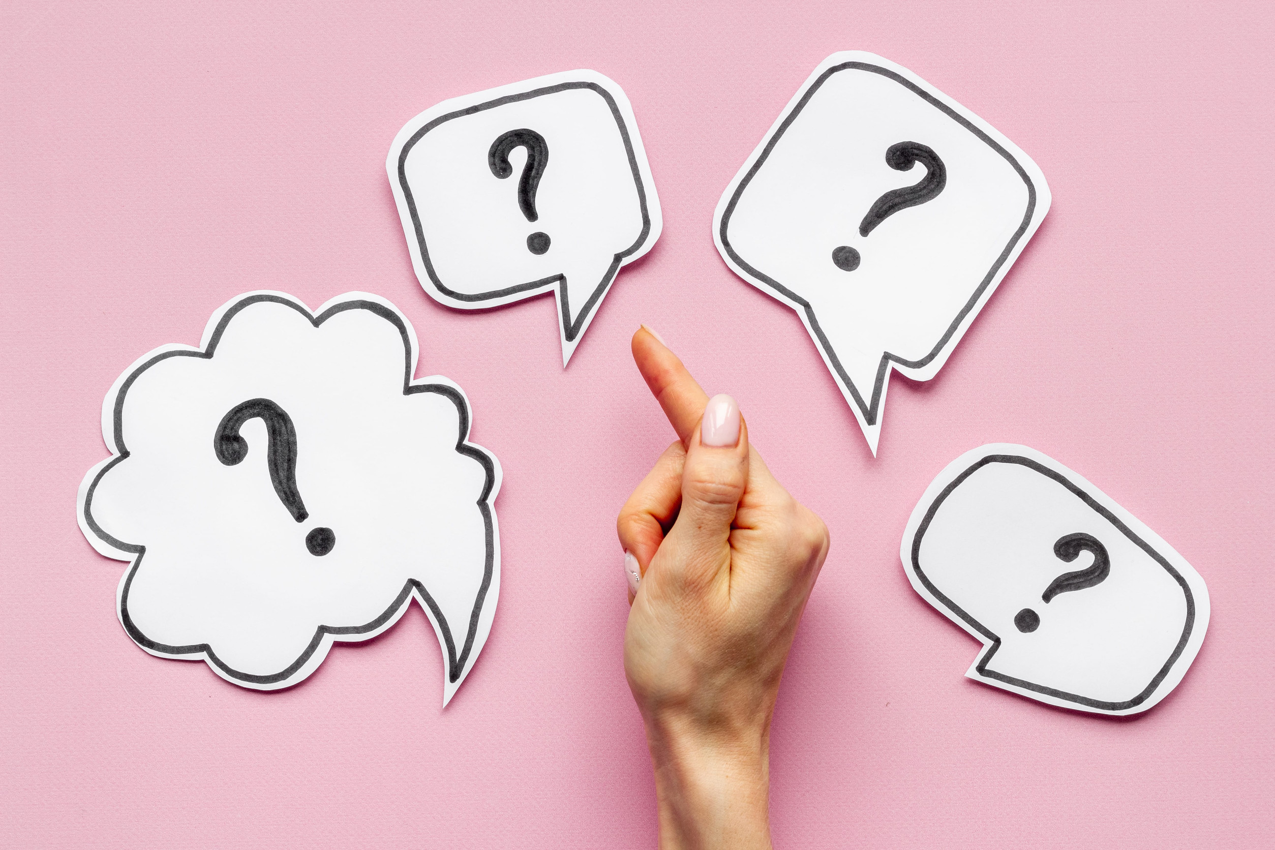 Hands point to white speech bubble cutouts with question marks on a pink background, symbolizing questions or choices.