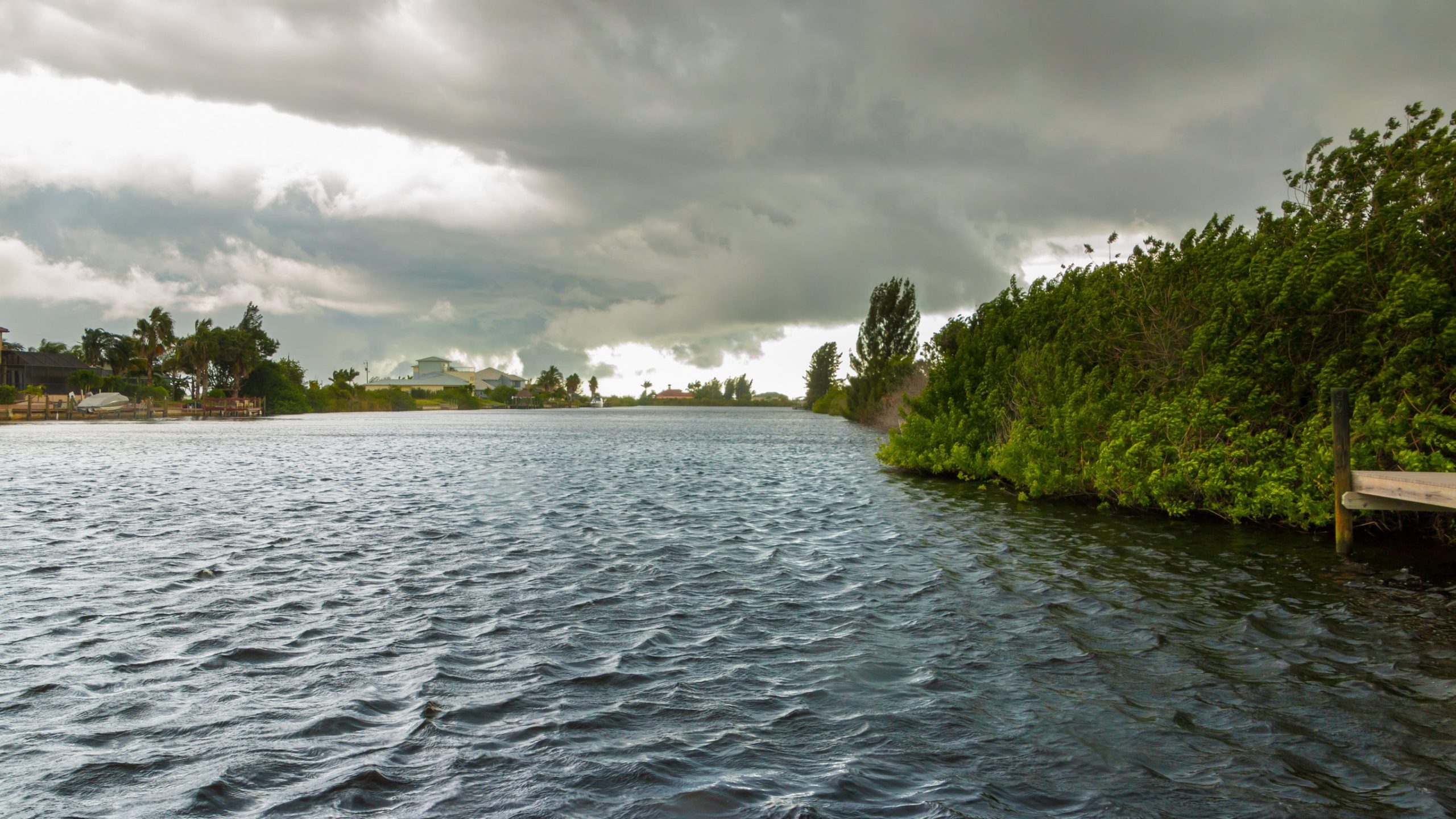 A wide, rippling river flows under a cloudy, overcast sky, with green bushes and trees along the right bank and houses visible in the distance on the left—homes that stand as a valuable investment thanks to their thoughtful placement and natural storm damage protection provided by the surrounding landscape.