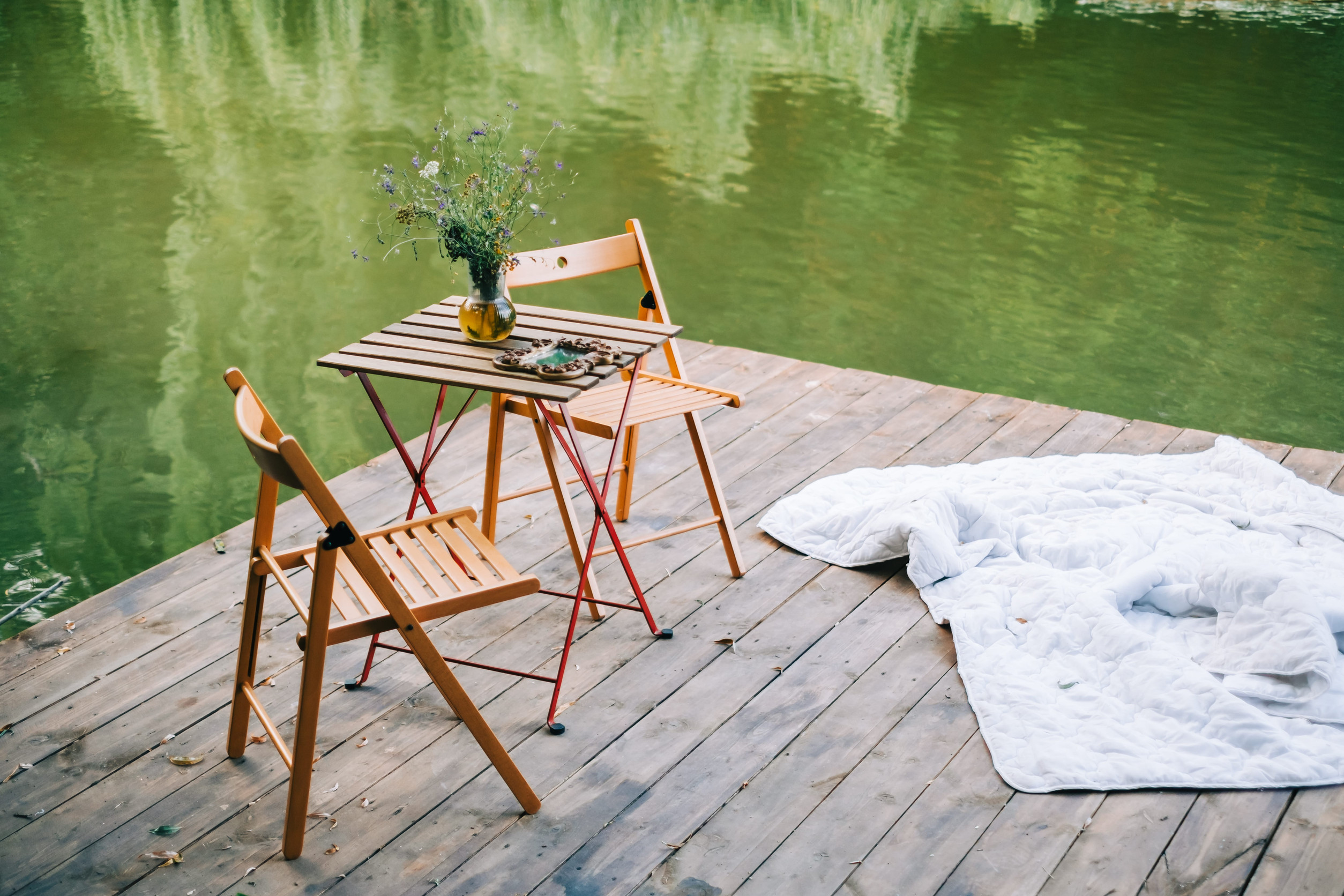 A small wooden table with two chairs sits on a dock by a green lake, offering inspiration for outdoor dock ideas. The table holds a vase of wildflowers and snacks, creating a charming outdoor living space. A white blanket lies casually on the wooden boards nearby.